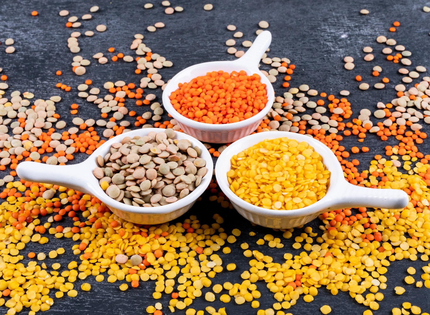 Different lentils in a mini white spice bowls on a black stone background. side view.